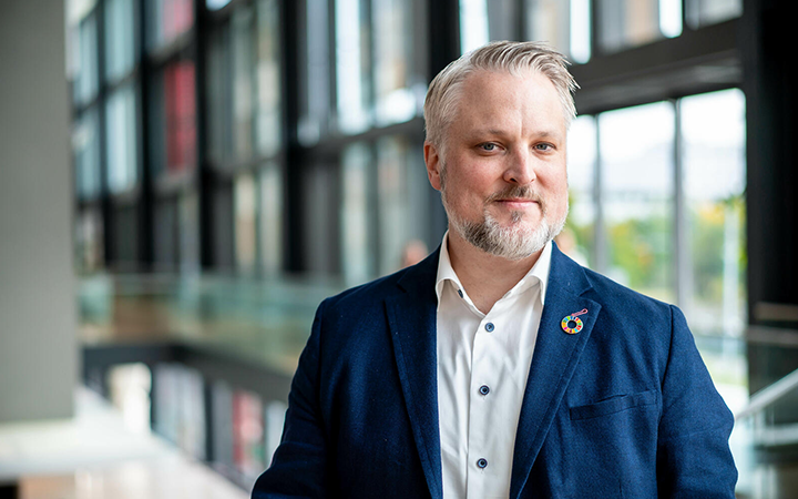 Philip Ripman i businesskläder står inomhus, omgiven av glasväggar. Han bär en mörkblå kavaj och en vit skjorta, med en färgglad pin på slaget