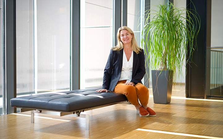 Kvinna i kavaj och orange byxor sitter på en modern bänk i ett ljust kontor med stora fönster och en grön växt i bakgrunden