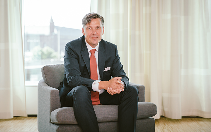 Per Wennberg i kostym sitter i en fåtölj framför ett fönster, inomhusmiljö med gardiner och dagsljus