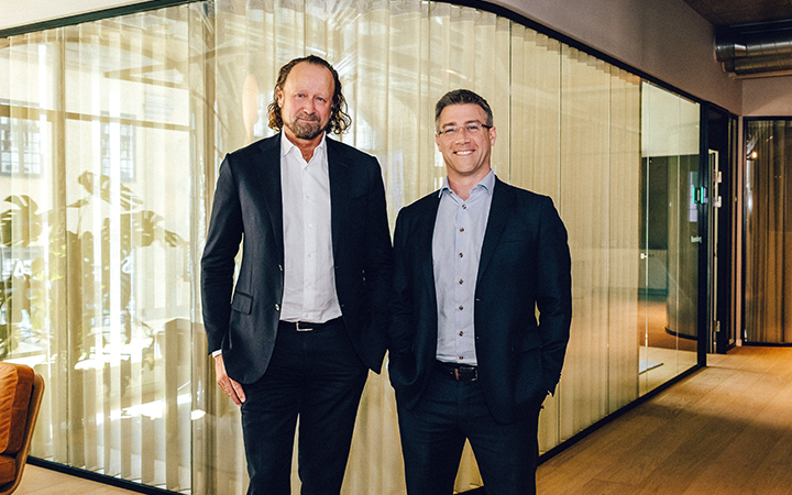 Två affärsmän i kostymer står i ett modernt kontor med glasväggar