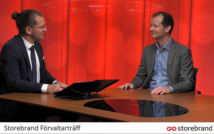 Två män i kostymer har ett samtal vid ett konferensbord under Storebrands förvaltarmöte, med röd bakgrund