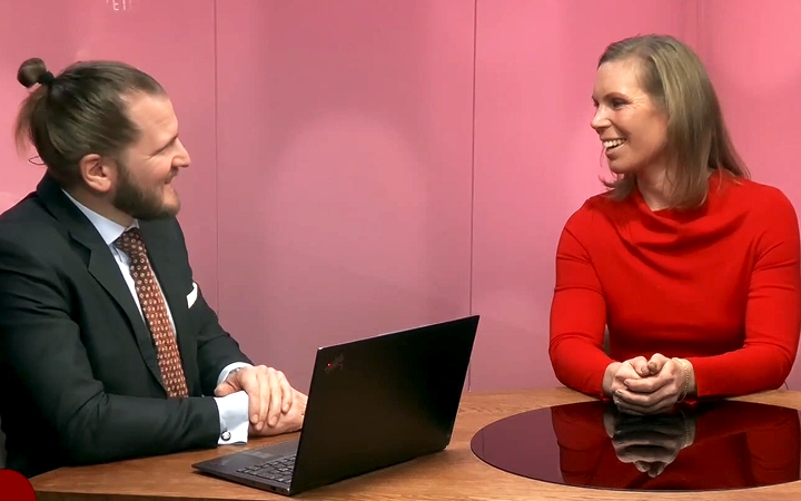 Man i kostym och kvinna i röd tröja diskuterar vid ett bord med en bärbar dator, mot en rosa bakgrund