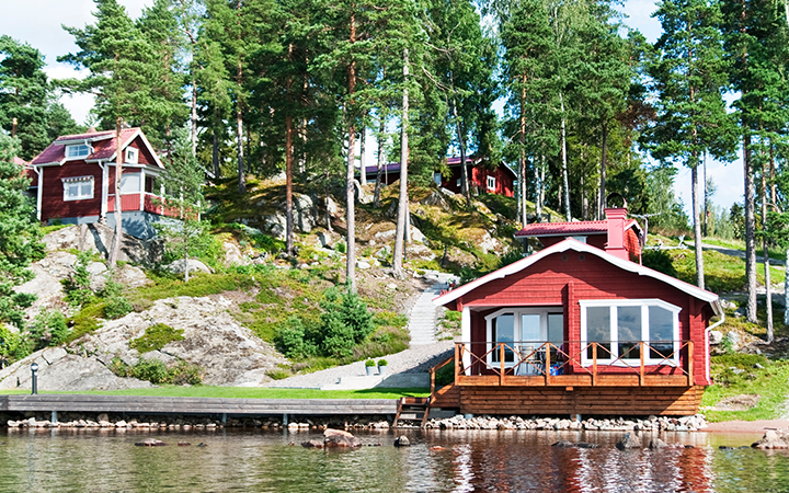 Röd stuga vid sjön i svensk skogsmiljö med tallar och gröna kullar