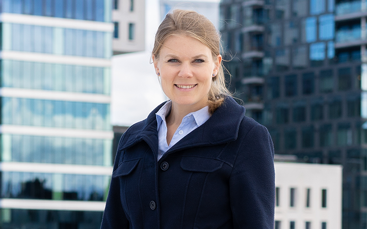 Sunniva Bratti mörk kappa i urban miljö med moderna byggnader i bakgrunden