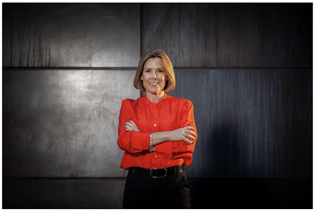 Kvinna i röd blus står mot en mörk bakgrund med armarna i kors och ler