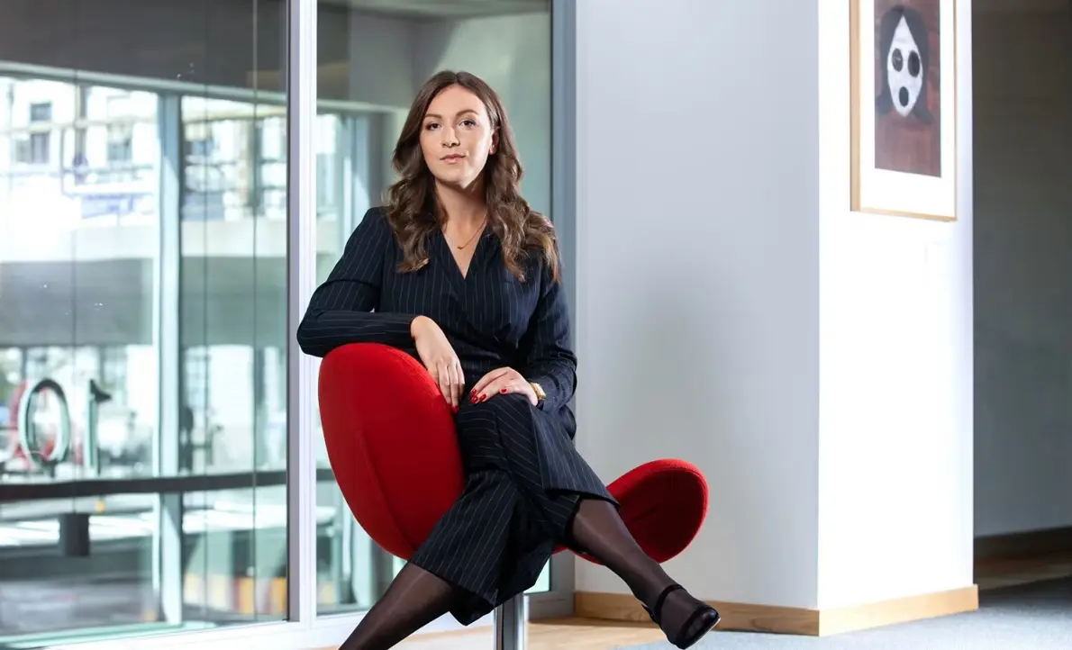 Kvinna i affärsdräkt sitter på en röd stol i ett modernt kontor med stora glasfönster i bakgrunden