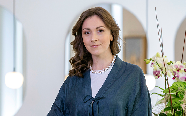 Kvinna i blå kappa med långt, brunt lockigt hår, står i ett modernt rum med vita väggar och orkidéer i bakgrunden