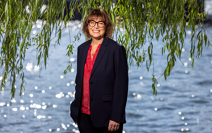 Kvinna i mörk kostym och röd blus framför en sjö, omgiven av hängande trädgrenar i solljus