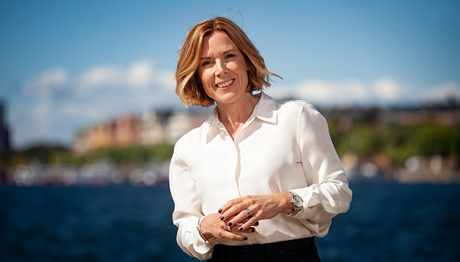 Kvinna i vit skjorta står framför en sjö med stadsmiljö i bakgrunden, en solig dag