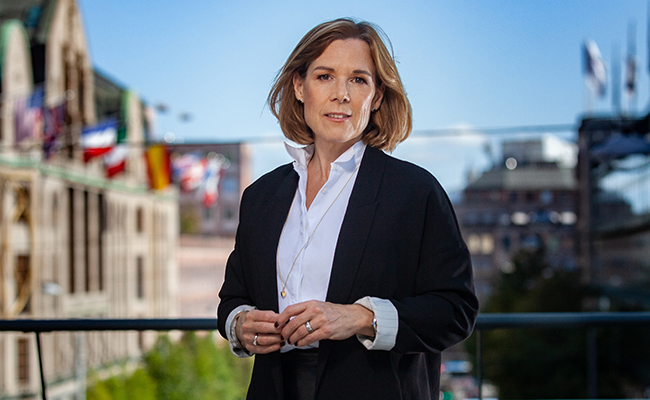 Kvinna i kostym står utomhus med stadsmiljö i bakgrunden, flaggor synliga