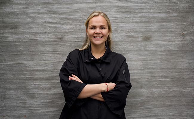 Ung kvinna med ljust hår i svart skjorta står framför en grå vägg och ler med armarna i kors