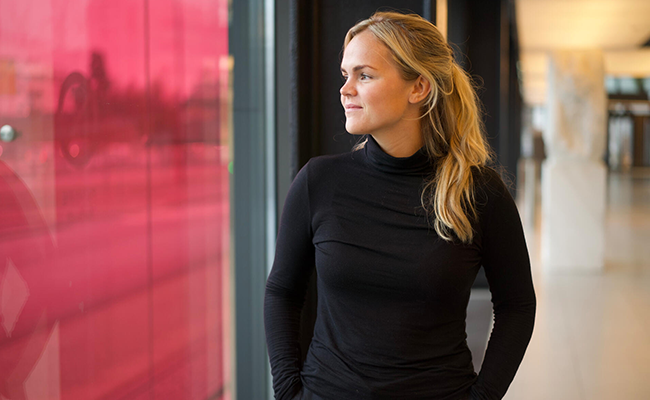 Kvinna i svart tröja står inomhus och tittar ut genom ett fönster med rosa bakgrund