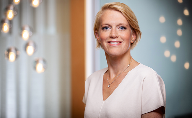 Kvinna med kort blont hår i vit blus, ler framför en modern inredning med ljusa glödlampor i bakgrunden