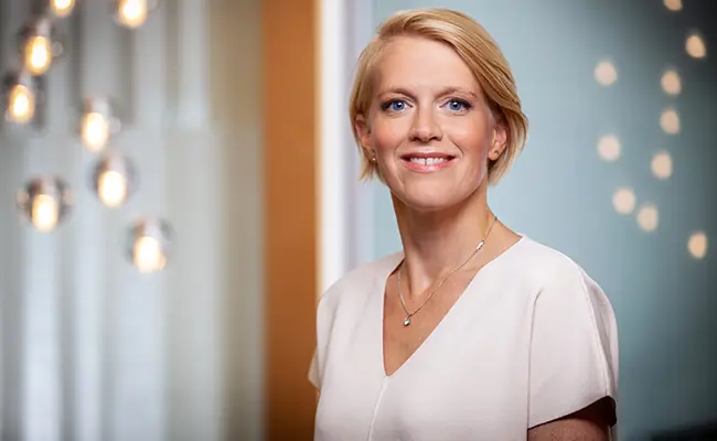 Kvinna med kort blont hår i vit blus, ler framför en modern inredning med ljusa glödlampor i bakgrunden