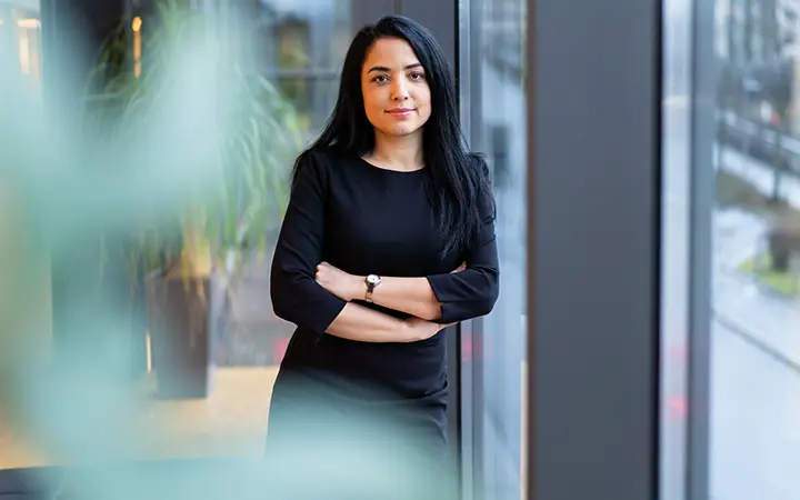 Kvinna i svart klänning står vid ett fönster, ser självsäker ut, ljus och modern kontorsmiljö i bakgrunden