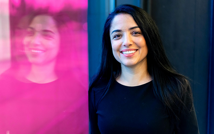 Kvinna med långt mörkt hår ler framför en rosa vägg, iklädd en svart tröja