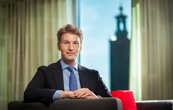 En man i kostym sitter i en soffa med Stockholms stadshus i bakgrunden, soligt dagsljus genom fönstret