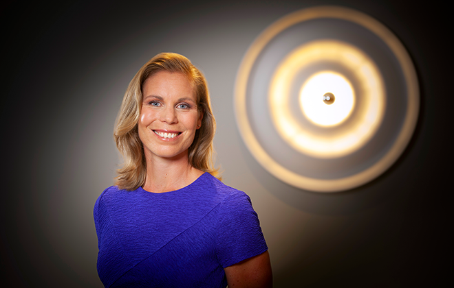 Anna Jönsson i blå klänning med kort blont hår ler mot kameran, med ett cirkulärt konstverk i bakgrunden