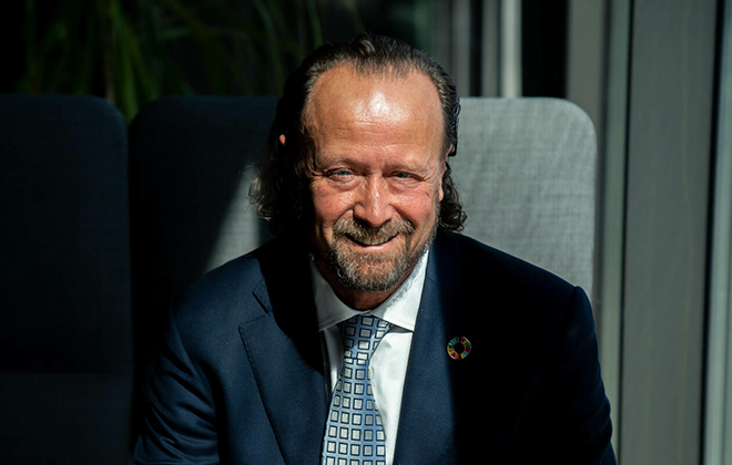 En man i kostym och slips sitter på en soffa. Bakgrunden är delvis belyst av solljus som strålar in genom ett fönster