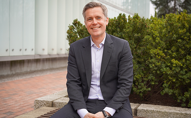 En leende man i kostym sitter utomhus på en bänk framför gröna buskar och en modern byggnad