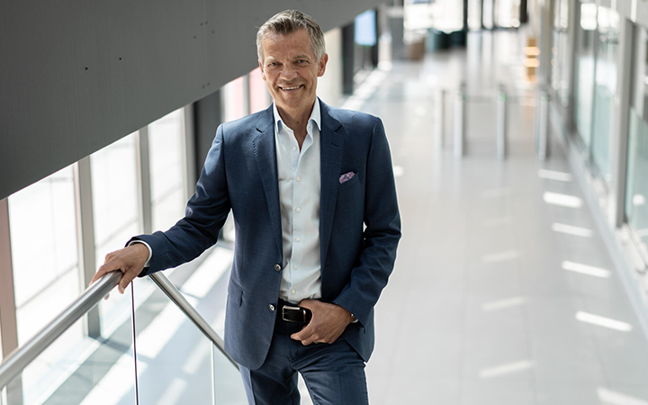 Äldre man i elegant kostym står i en modern korridor med glasväggar, ser självsäker ut och håller handen på ledstången