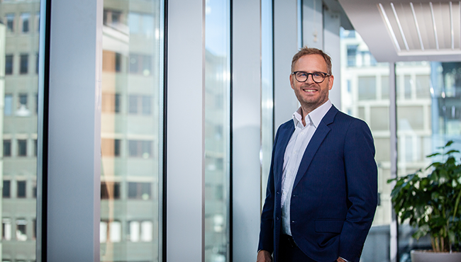 Man i kostym står vid fönster på kontor, med urbana byggnader i bakgrunden