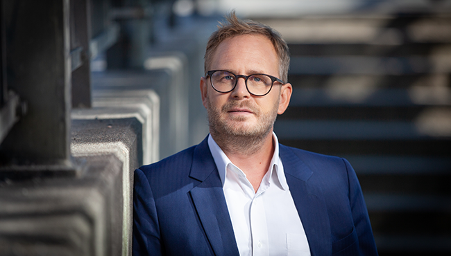 Man med glasögon och kostym står utomhus vid en trappa, sommarklädd i soligt väder