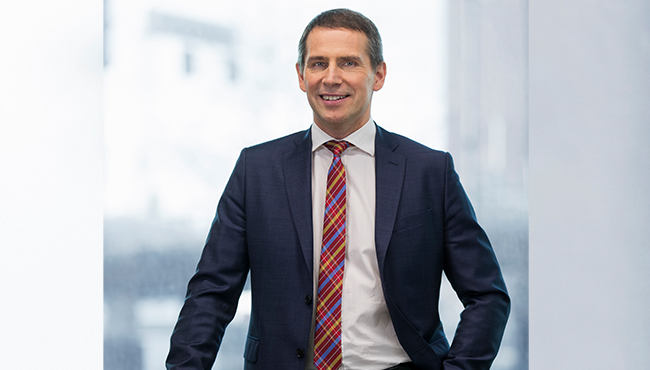 En leende man i mörk kostym och rutig slips står framför ett fönster i ett kontor