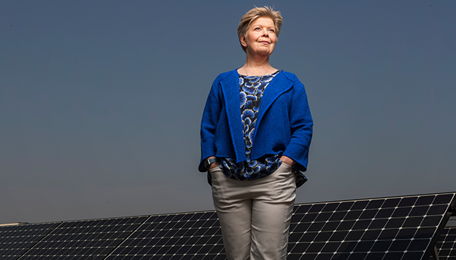 Kvinna står framför solpaneler under en klarblå himmel, iklädd blå jacka och mönstrad blus