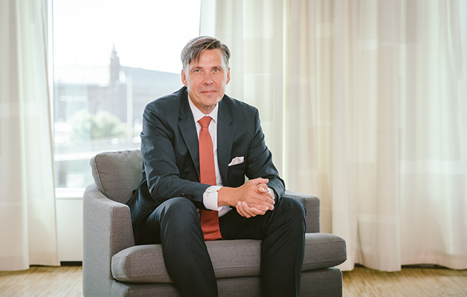 Man i kostym sitter i en fåtölj framför ett solbelyst fönster, med gardiner i bakgrunden