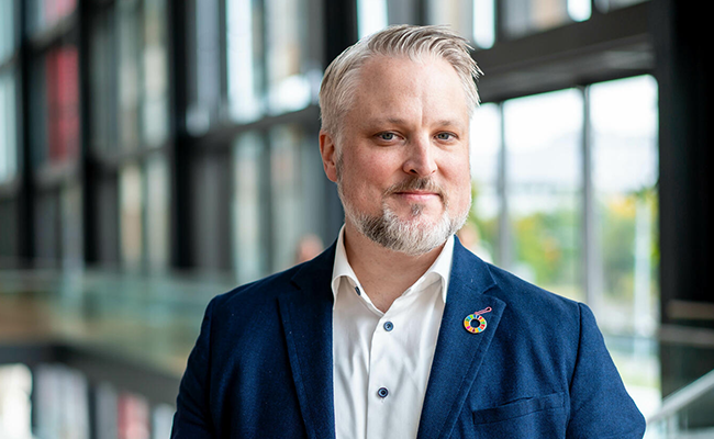 Man i blå kostym med vit skjorta står inomhus framför stora fönster