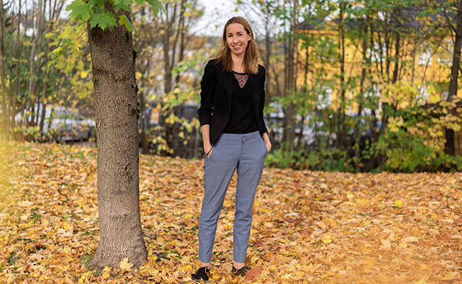Kvinna i svart topp och grå byxor står i höstlandskap med löv på marken