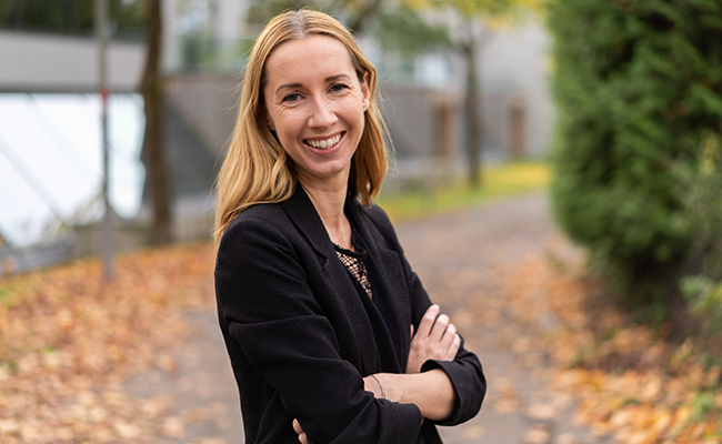 Kvinna i svart kavaj med ett leende, står på en höstig gångväg med gulnade löv