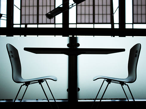 Två tomma stolar bredvid ett bord i ett modernt kontorsutrymme, silhuett mot ett stort fönster i bakgrunden