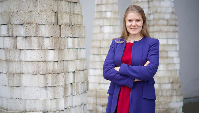 Kvinna i lila kappa och röd klänning står med armarna i kors framför vita tegelkolonner