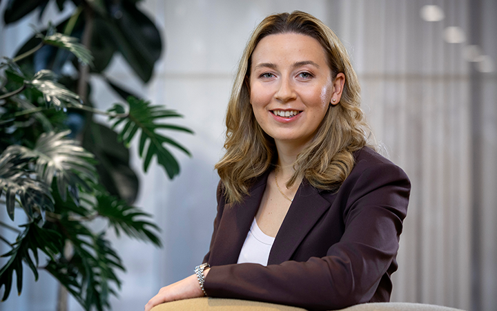 Kvinna i brun kavaj sitter leende vid ett skrivbord med en grön växt i bakgrunden