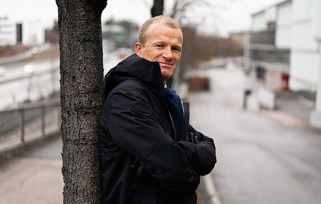 En person i mörk jacka lutar mot ett träd på en stadsgata, vinterdag med molnigt väder i bakgrunden