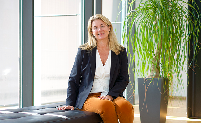 Kvinna i formell klädsel sitter på en soffa i ett modernt kontor med stora fönster och en grön växt i bakgrunden