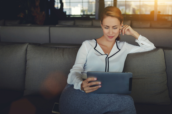 Kvinna i vit blus sitter bekvämt på soffa och läser på en surfplatta i modernt vardagsrum