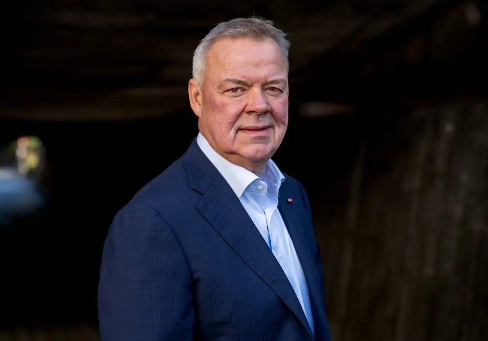 Odd Arild i mörk kostym står utomhus i skuggan, leende lätt mot kameran