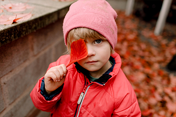 Litet barn i rosa jacka och mössa håller upp ett höstlöv framför ögat, omgiven av fallna röda löv