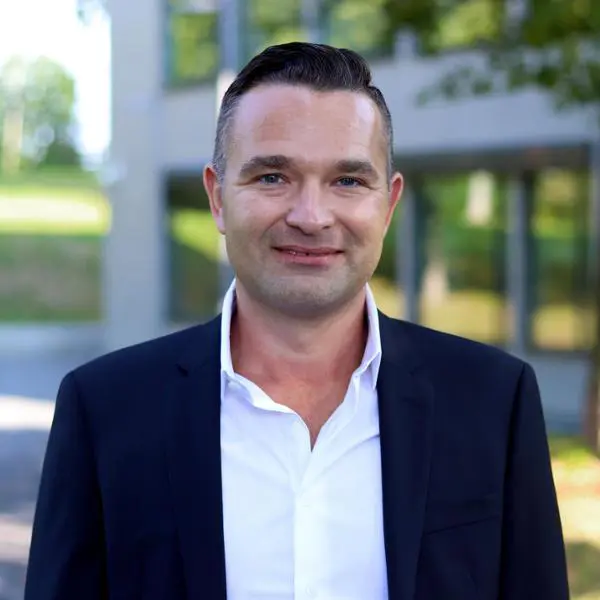 Man i mörk kostym och vit skjorta utomhus framför en modern byggnad