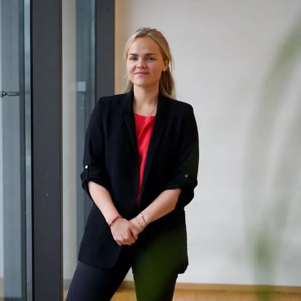 Kvinna i svart kavaj och röd topp står vid ett fönster i en modern interiör