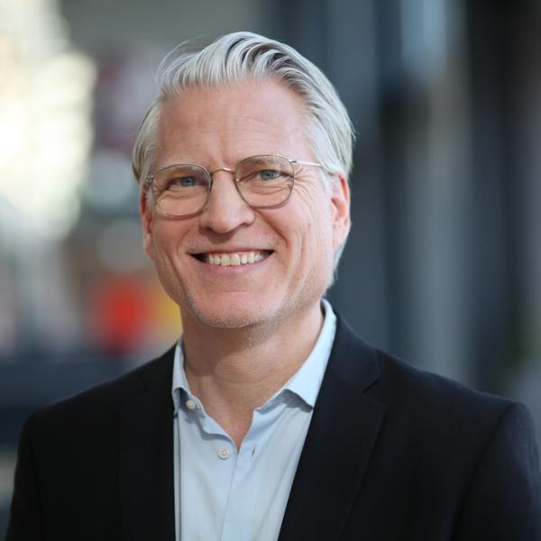 Äldre man med glasögon och grått hår i en mörk kostymjacka, leende i en inomhusmiljö