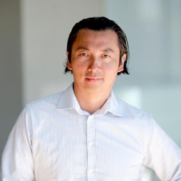 En person i vit skjorta står i en ljus miljö, tittar in i kameran med ett neutralt ansiktsuttryck