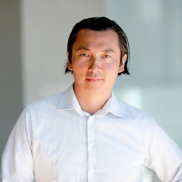 En person i vit skjorta står i en ljus miljö, tittar in i kameran med ett neutralt ansiktsuttryck