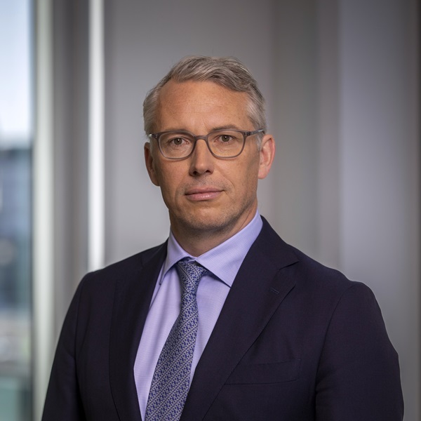 En person i kostym med glasögon och ljus skjorta, står framför ett kontorsfönster