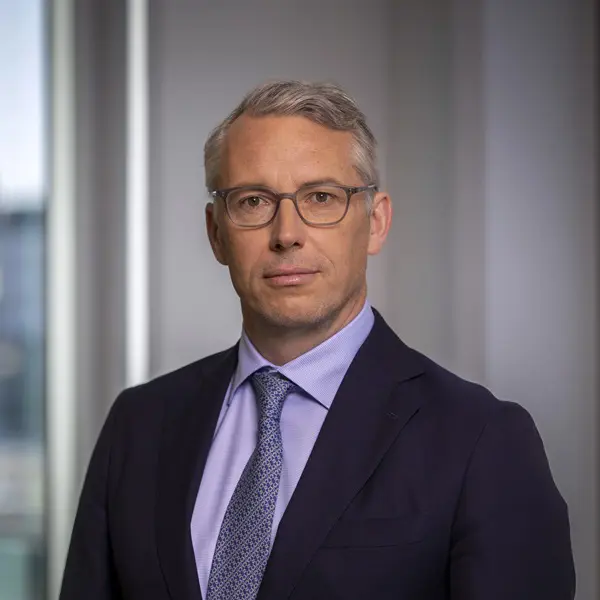 En person i kostym med glasögon och ljus skjorta, står framför ett kontorsfönster